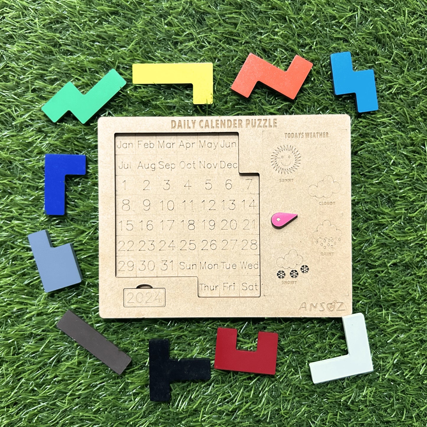 A wooden daily calendar puzzle toy arranged on artificial green grass, surrounded by colorful L-shaped wooden pieces. The central wooden board shows a numbered calendar grid with days of the week and small printed illustrations.