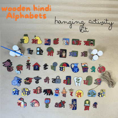 A wooden Hindi alphabet hanging activity kit arranged on a beige textured background, with orange text reading 'wooden hindi Alphabets' and black script text reading 'hanging activity kit.' The kit includes many colorful wooden Hindi letters shaped like everyday objects, small hanging pegs, a paintbrush, two small bottles, and a string.