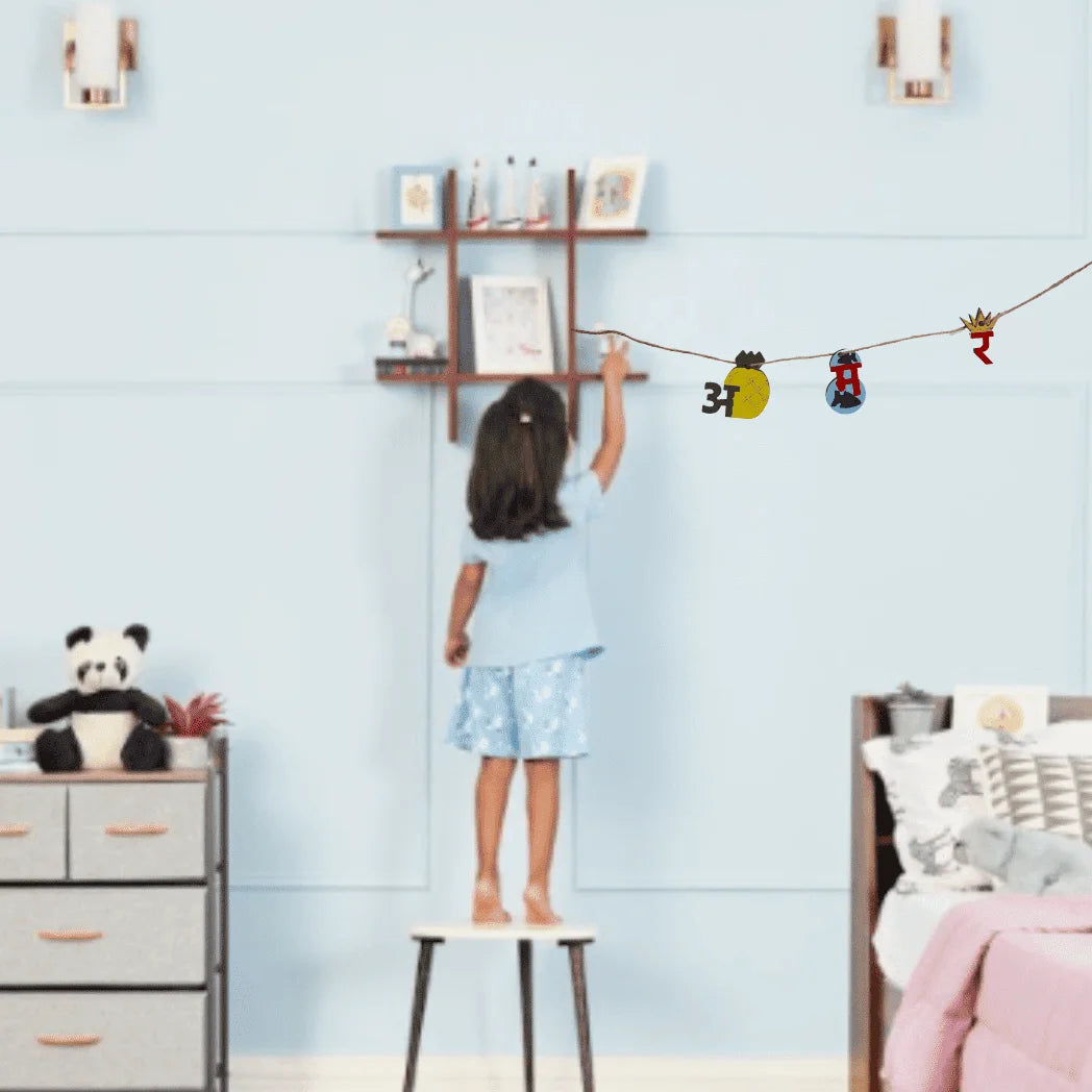 A child stands on a small stool in a bedroom, reaching toward wall decals hanging on a string. The room has light blue walls, a wooden shelf with framed pictures, a panda plush on a dresser, and a bed with pink bedding.