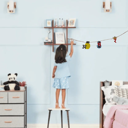 A child stands on a small stool in a bedroom, reaching toward wall decals hanging on a string. The room has light blue walls, a wooden shelf with framed pictures, a panda plush on a dresser, and a bed with pink bedding.