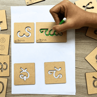 Child's hand tracing Telugu alphabet letters on wooden stencils with a green sketch pen.