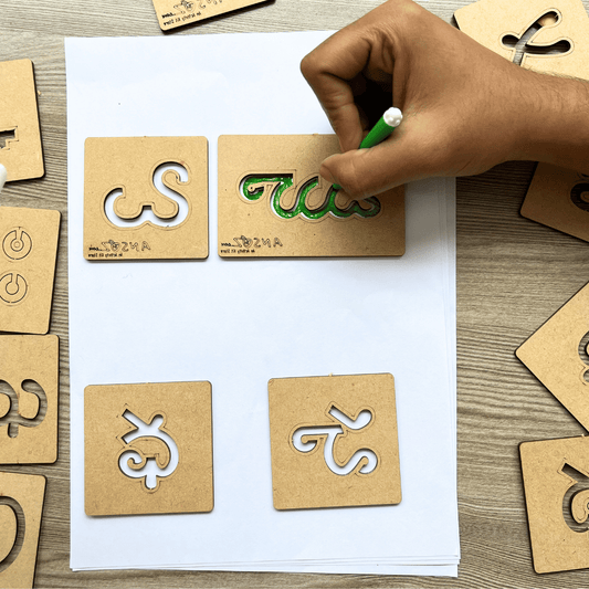 Child's hand tracing Telugu alphabet letters on wooden stencils with a green sketch pen.