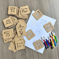 A set of wooden Hindi learning stencil board arranged on a light wood surface, with a white worksheet, colored markers, and additional scattered cards.