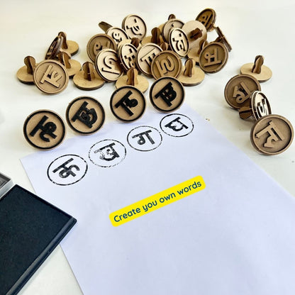 A collection of wooden Hindi alphabet stamps arranged around a sheet of paper, showing stamped Hindi letters. An ink pad is placed beside the paper, demonstrating how children can create their own Hindi words and practice Hindi Swar and Vyanjan through hands-on learning.