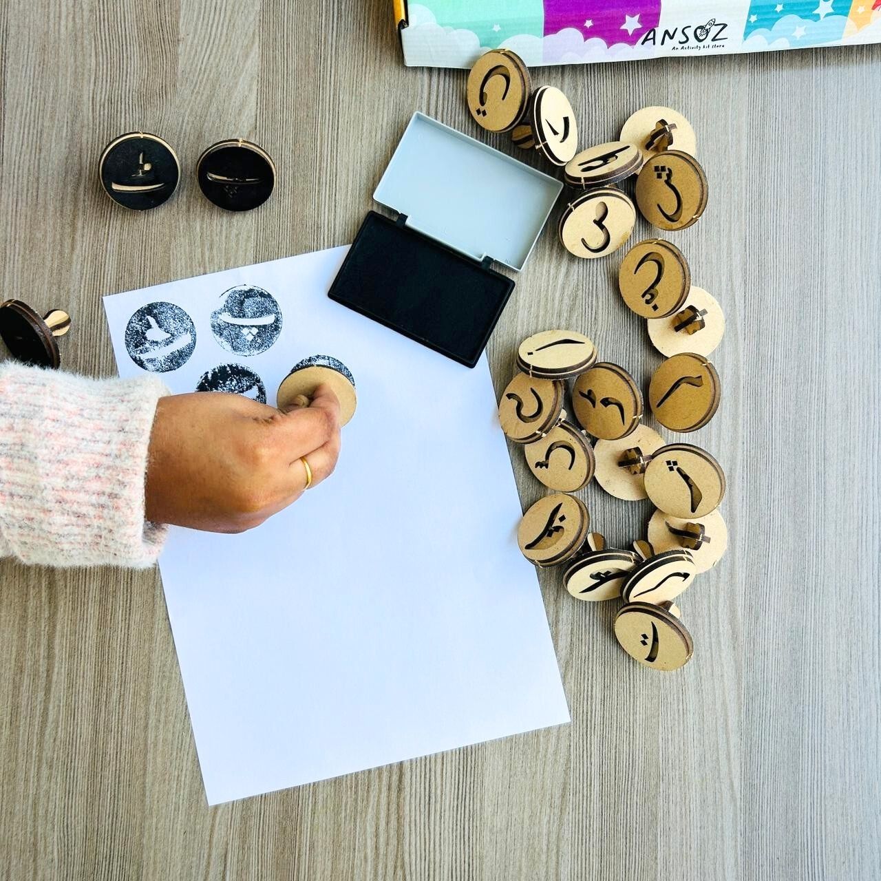 The child uses an Urdu alphabet stamp on paper with a stamp kit and ink pad for educational games.
