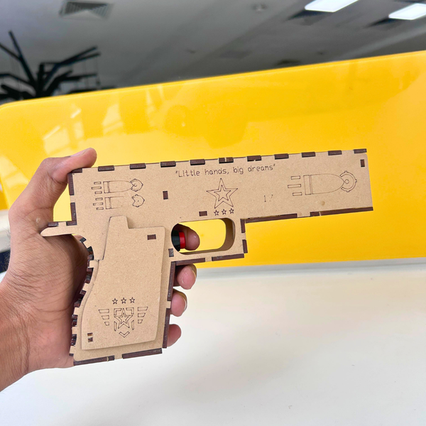 Hand holding DIY wooden laser gun science kit with laser-cut design and yellow background for STEM learning