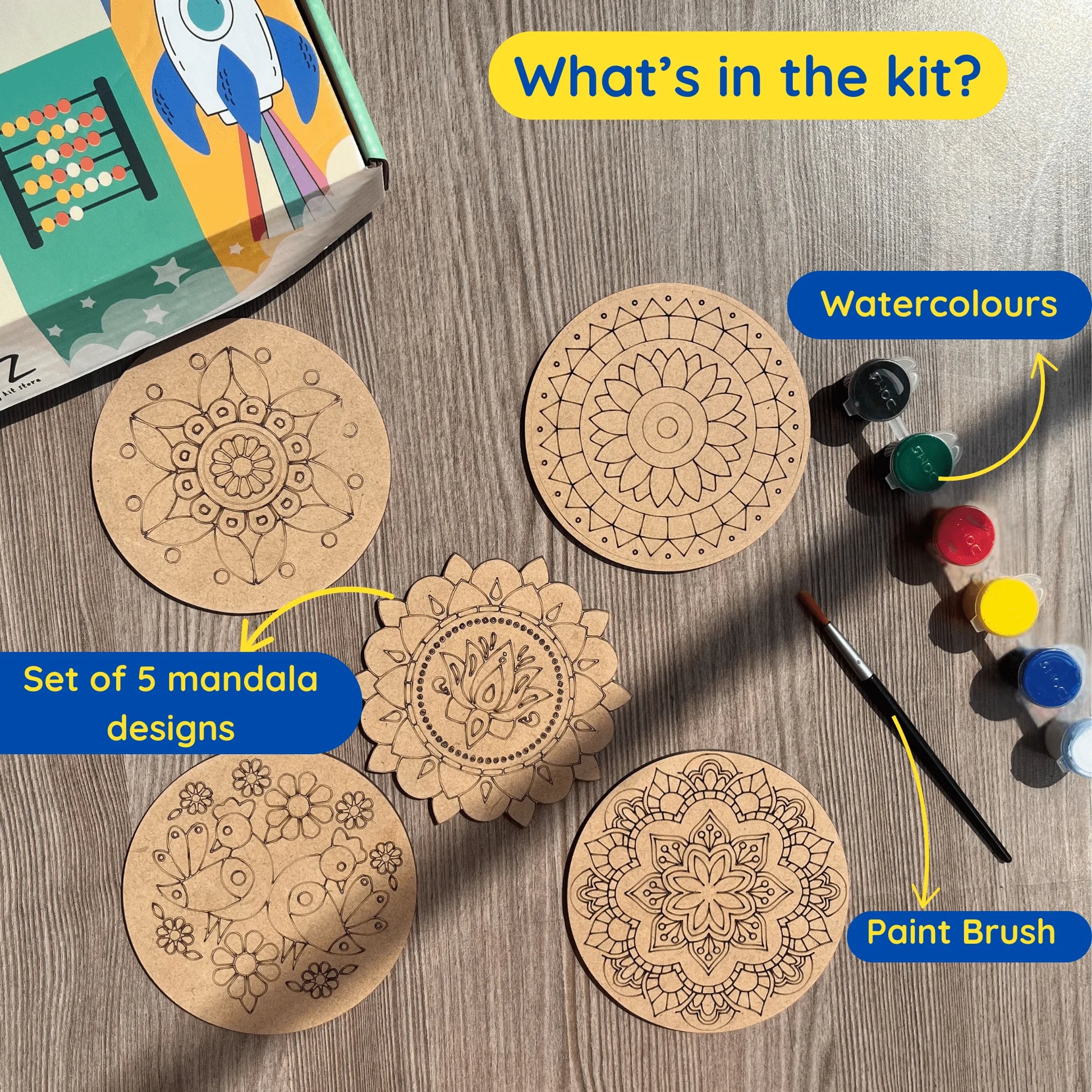 A flat-lay product presentation of a mandala painting kit on a gray wood-textured background. The kit includes five circular mandala templates with hand-drawn black line art, a watercolor palette with five colors (black, green, red, yellow, blue), and a paint brush.