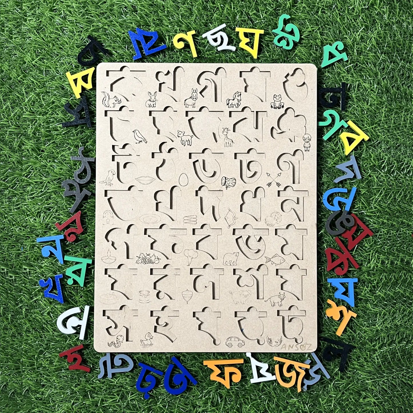 A wooden Bengali alphabet learning board with cutout letters and small illustrations, surrounded by colorful 3D Bengali letters arranged on artificial grass.