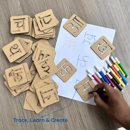 A hand is using a blue marker to trace a wooden Hindi letter card on a white sheet of paper, surrounded by other wooden Hindi letter cards and colored drawing pens on a gray wood-textured surface. Blue text at the bottom reads 'Trace, Learn & Create'.