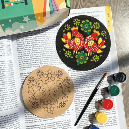 Two round wooden coasters with laser-engraved floral designs, shown next to a paintbrush and small paint pots on top of an open book. One coaster is painted black with colorful birds and flowers, and the other is natural wood.