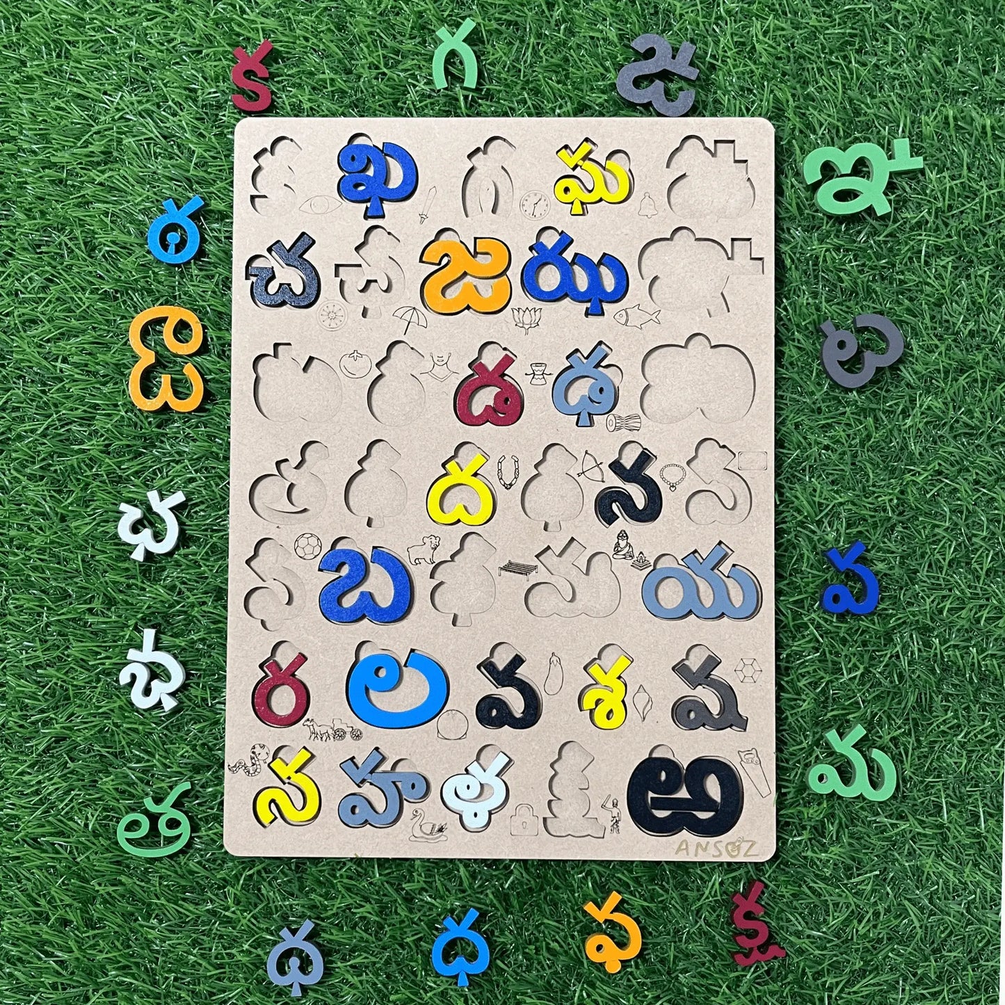 A wooden puzzle board featuring cutout shapes and colorful Telugu letters arranged around it on a green grass-like surface.