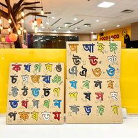 Three wooden Bengali alphabet learning boards with colorful painted letters, displayed on a white surface in front of a yellow wall inside a store.