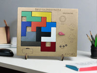 A wooden daily calendar puzzle stand on a white desk, featuring colorful sliding puzzle blocks and weather icons. The product is branded with the name 'ANSOZ' on the bottom right.