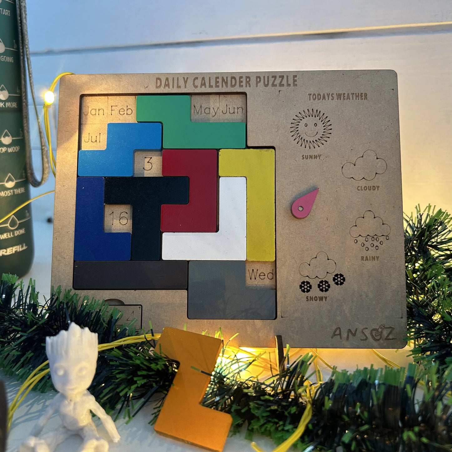A wooden daily calendar puzzle board with colorful interlocking puzzle pieces and weather icons, styled with green garland and warm string lights.