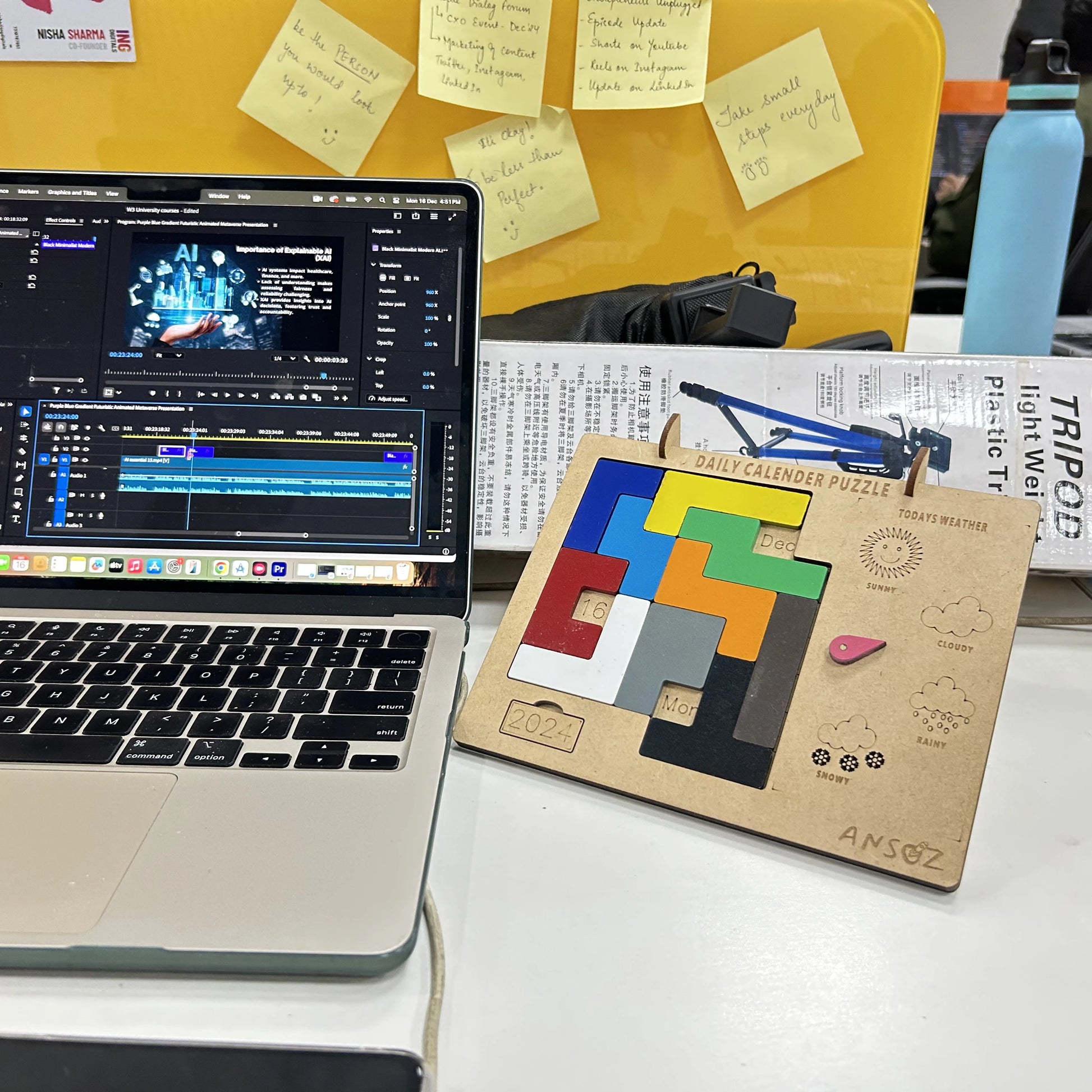 A wooden daily calendar puzzle board with colorful geometric pieces and a small pink eraser sits on a desk next to an open laptop showing video editing software. A yellow board with sticky notes and a blue water bottle are visible in the background.