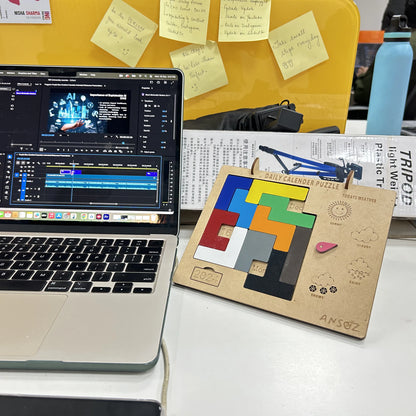 A wooden daily calendar puzzle board with colorful geometric pieces and a small pink eraser sits on a desk next to an open laptop showing video editing software. A yellow board with sticky notes and a blue water bottle are visible in the background.