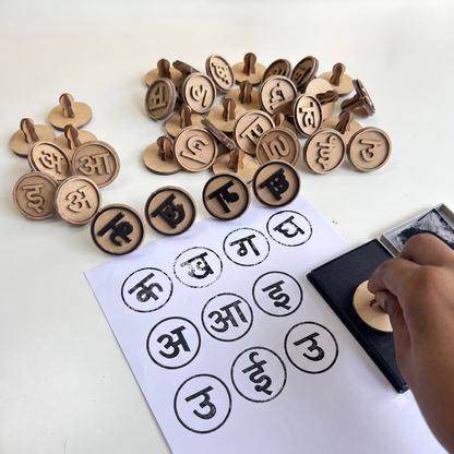 A Hindi Vowel and Consonant Stamp Kit displayed on a white surface, featuring wooden Hindi Swar and Vyanjan stamps, an ink pad, and a sheet of paper with stamped Hindi letters. A hand is using a stamp to print letters, demonstrating a fun and interactive Hindi learning activity for kids. The educational kit helps children recognize Hindi alphabet letters, practice word formation, improve fine motor skills, and develop early Hindi language learning through hands-on play.