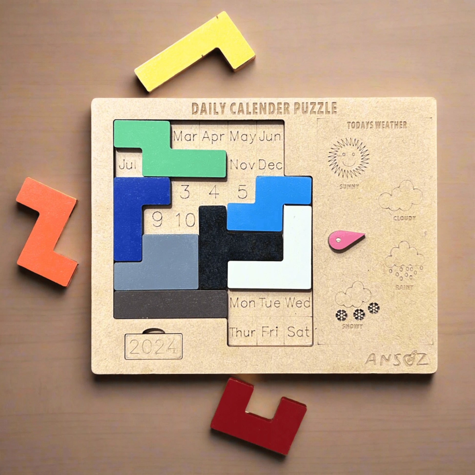 A wooden daily calendar puzzle for 2024 shown from above on a brown surface, with several colorful puzzle pieces placed around it. The main board is light natural wood with engraved weather icons and days of the week, and the movable pieces include green, blue, black, gray, white, pink, yellow, orange, and red.
